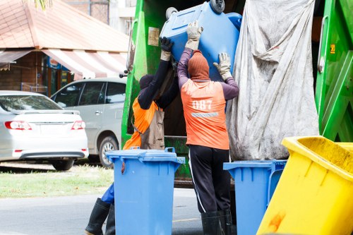 Two waste collection workers are loading a large blue plastic caddy from a green refuse collection vehicle parked on a residential street, with one worker wearing an orange high-visibility vest and black gloves, and the other wearing a purple hooded jacket and gloves. The vehicle's rear end is open, revealing a grey tarp covering part of the loading area. In the foreground, two empty blue bins and a yellow waste bin are visible on the pavement, while in the background, parked cars and a house with a cream-colored brick exterior and a striped awning can be seen, suggesting a suburban area within or near West Byfleet. The scene is illuminated by natural daylight, indicating a typical working day for rubbish removal services provided by House Clearance West Byfleet, operating in the local postcode area.