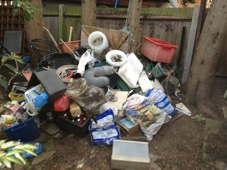 A large pile of mixed household waste and rubbish collected in a garden setting, featuring various discarded items including plastic bags, cardboard boxes, a black plastic crate, two white plastic pipes, a red plastic container, a grey PVC pipe, and a grey metal box. The rubbish is spread across the soil ground, surrounding wooden fence posts and a weathered wooden fence, with some items leaning against the fence or resting on the soil. The scene appears to be outdoors, likely in a residential backyard or garden area, suggesting a recent clearance or tidy-up task undertaken by a local rubbish removal service. The overall scene is cluttered and unorganized, with waste materials of different textures and colours, primarily including white, grey, black, and red, indicating items ready for collection or disposal. House Clearance West Byfleet often manages similar rubbish removal jobs in the West Byfleet area, ensuring efficient clearing of garden debris and household waste.