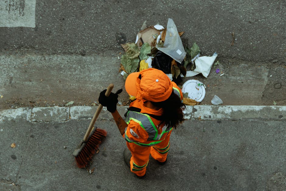 A green plastic skip filled with bags of gardening or landscaping materials, including red bags of flexible edging products, situated outdoors against a dark brick wall on a concrete surface. The skip