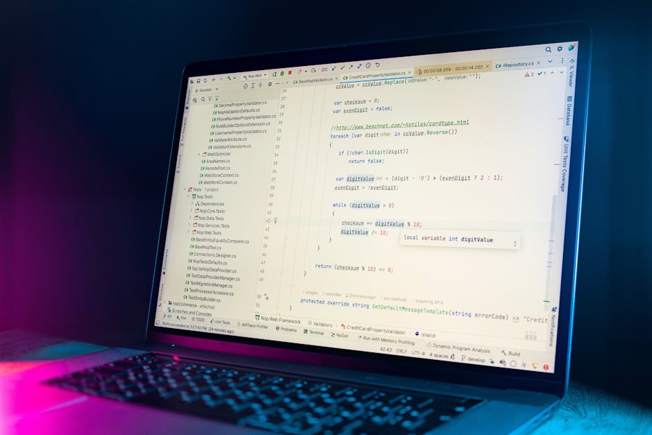 A close-up view of a person’s hands typing on a portable laptop placed on a dark, smooth surface. The laptop screen displays lines of colorful code on a dark background, with the interface showing a