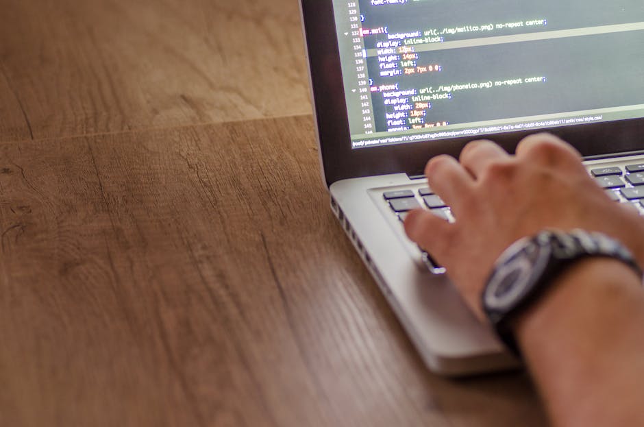 A person's right hand wearing a wristwatch is seen typing on a laptop keyboard placed on a wooden desk. The laptop screen displays lines of code with a dark background and multicolored text. The surfa