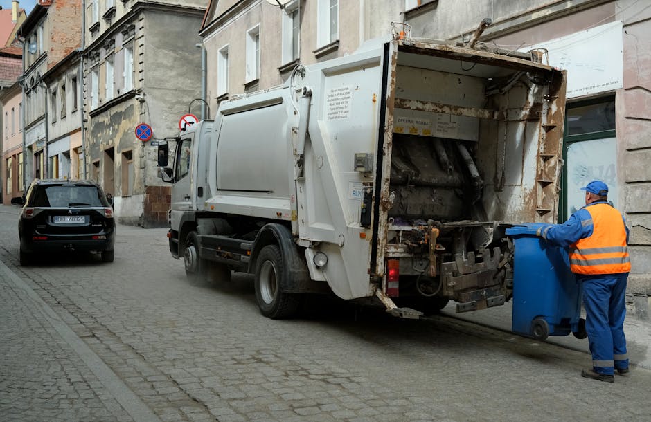 A row of large mobile waste bins aligned along a paved pathway beside a brick wall, with the bins positioned in a sequence featuring green, blue, and red lids, indicating different waste categories. T