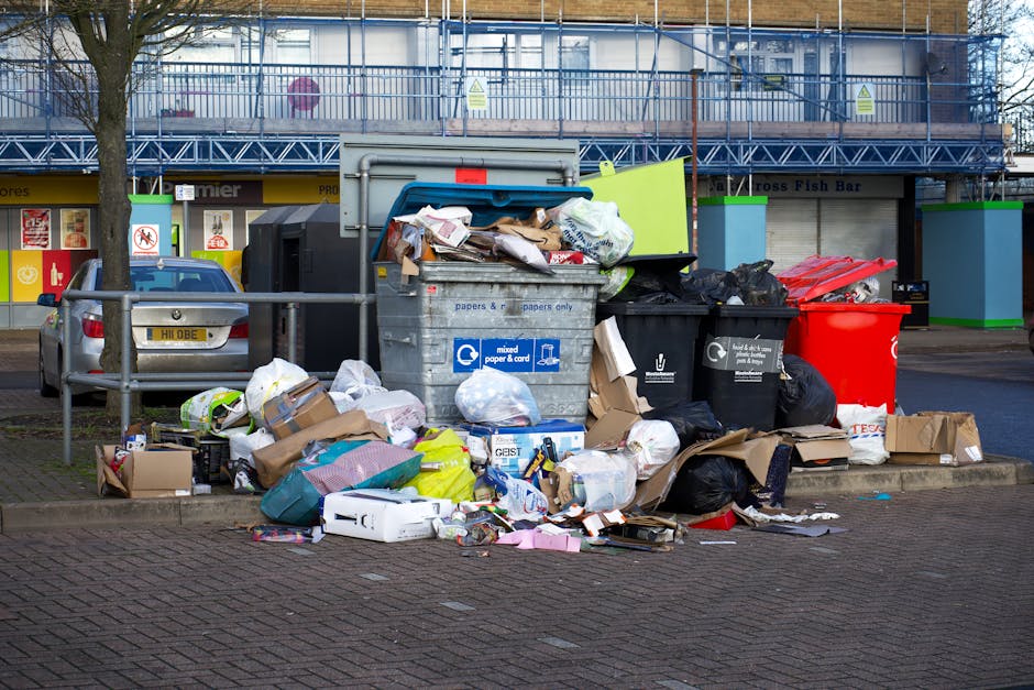 A small, older blue pickup truck parked on a street with its cargo area fully loaded with various discarded items, including large cardboard boxes, black plastic garbage bags, and miscellaneous debris